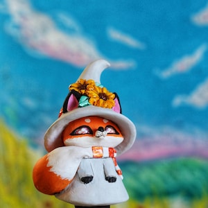 May include: A charming fox figure with orange fur, sporting a white witch's hat decorated with sunflowers. The fox is wearing a white cape and a scarf with orange stripes. The background features a blue sky and a green landscape.