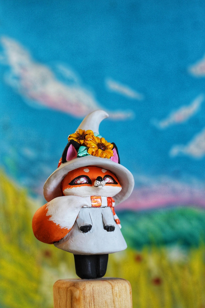 May include: A charming fox figure with orange fur, sporting a white witch's hat decorated with sunflowers. The fox is wearing a white cape and a scarf with orange stripes. The background features a blue sky and a green landscape.