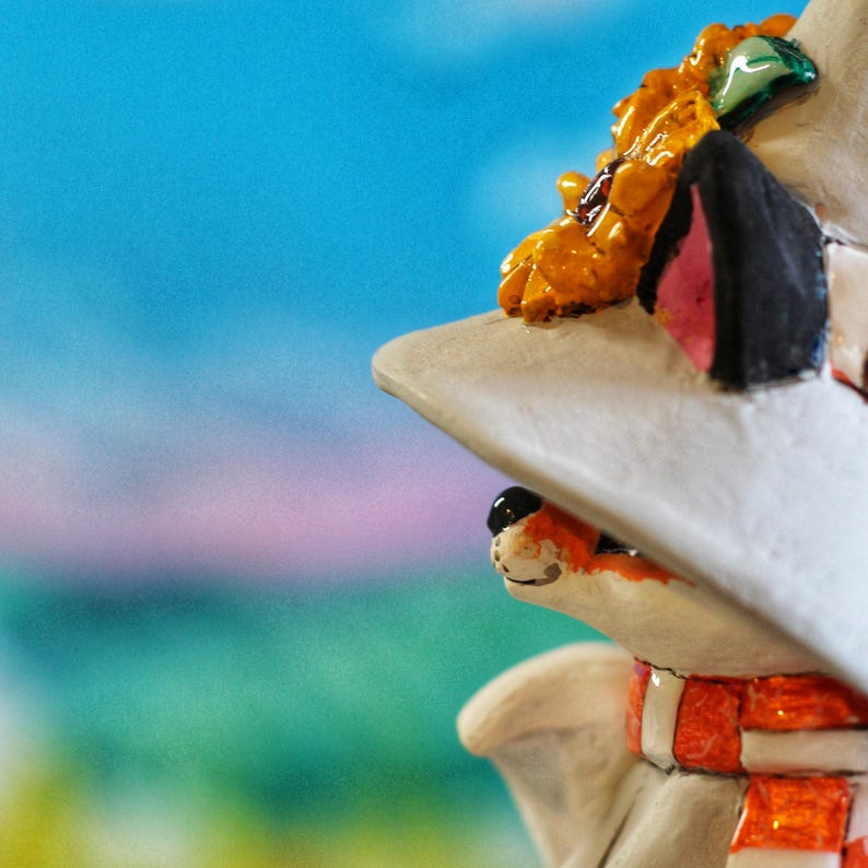 May include: A detailed figurine of a fox wearing a white hat, an orange and white scarf, and a decorative piece on top of the hat. The fox has black and pink ears and a detailed face. The background is a gradient of blue and pink.