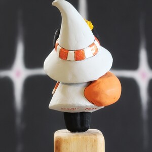 May include: A quirky figurine of a character wearing a white hat and an orange and white striped scarf. The figure has an orange rounded back and is mounted on a wooden block. The background is dark.