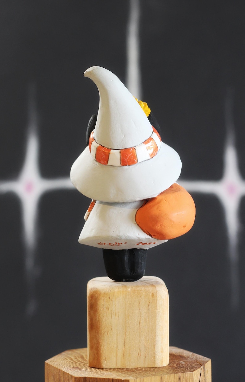 May include: A quirky figurine of a character wearing a white hat and an orange and white striped scarf. The figure has an orange rounded back and is mounted on a wooden block. The background is dark.