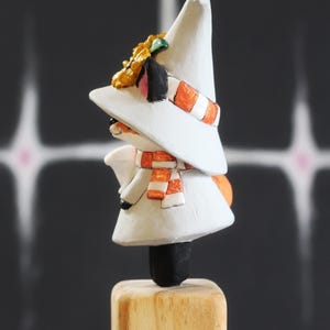 May include: A hand-painted figurine of a character wearing a white hat, scarf, and coat. The scarf has orange and white stripes. The figurine is mounted on a wooden base. The character has a black nose and ears, and a yellow flower on the hat.