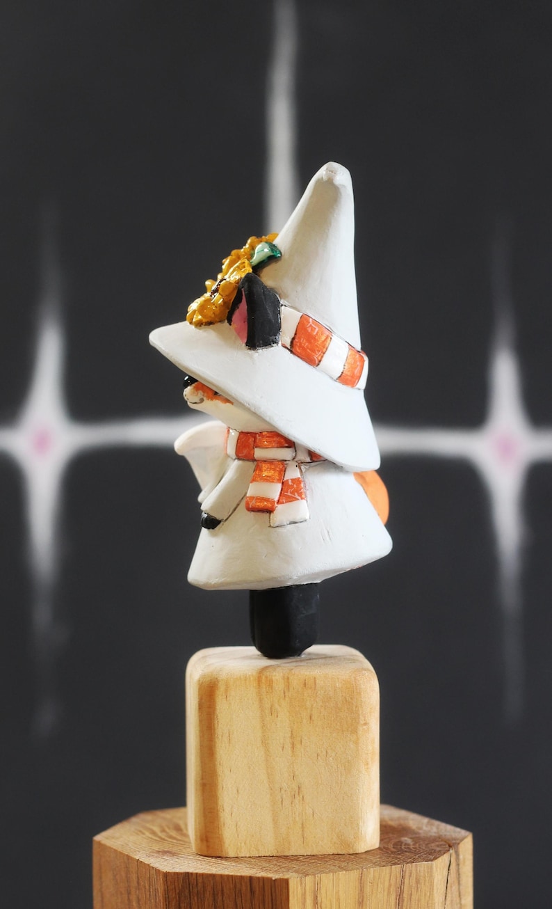 May include: A hand-painted figurine of a character wearing a white hat, scarf, and coat. The scarf has orange and white stripes. The figurine is mounted on a wooden base. The character has a black nose and ears, and a yellow flower on the hat.
