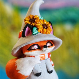 May include: A decorative fox figurine, wearing a grey witch's hat decorated with yellow sunflowers. The fox has orange fur, a white scarf, and a grey outfit. A charming, whimsical item.