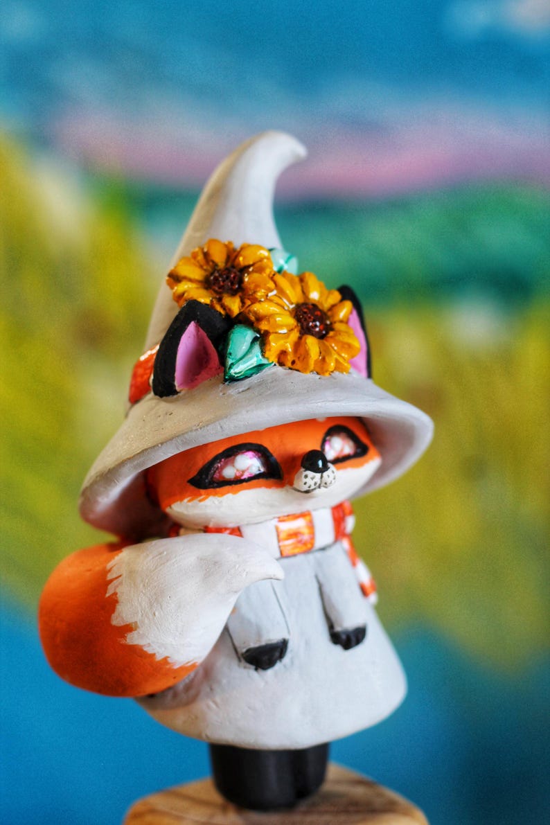 May include: A decorative fox figurine, wearing a grey witch's hat decorated with yellow sunflowers. The fox has orange fur, a white scarf, and a grey outfit. A charming, whimsical item.