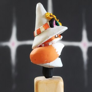 May include: A quirky wooden fox ornament. The fox is depicted wearing a white hat adorned with a sunflower and an orange and white striped band. The fox has an orange body and is mounted on a wooden base. The background is dark with white streaks.