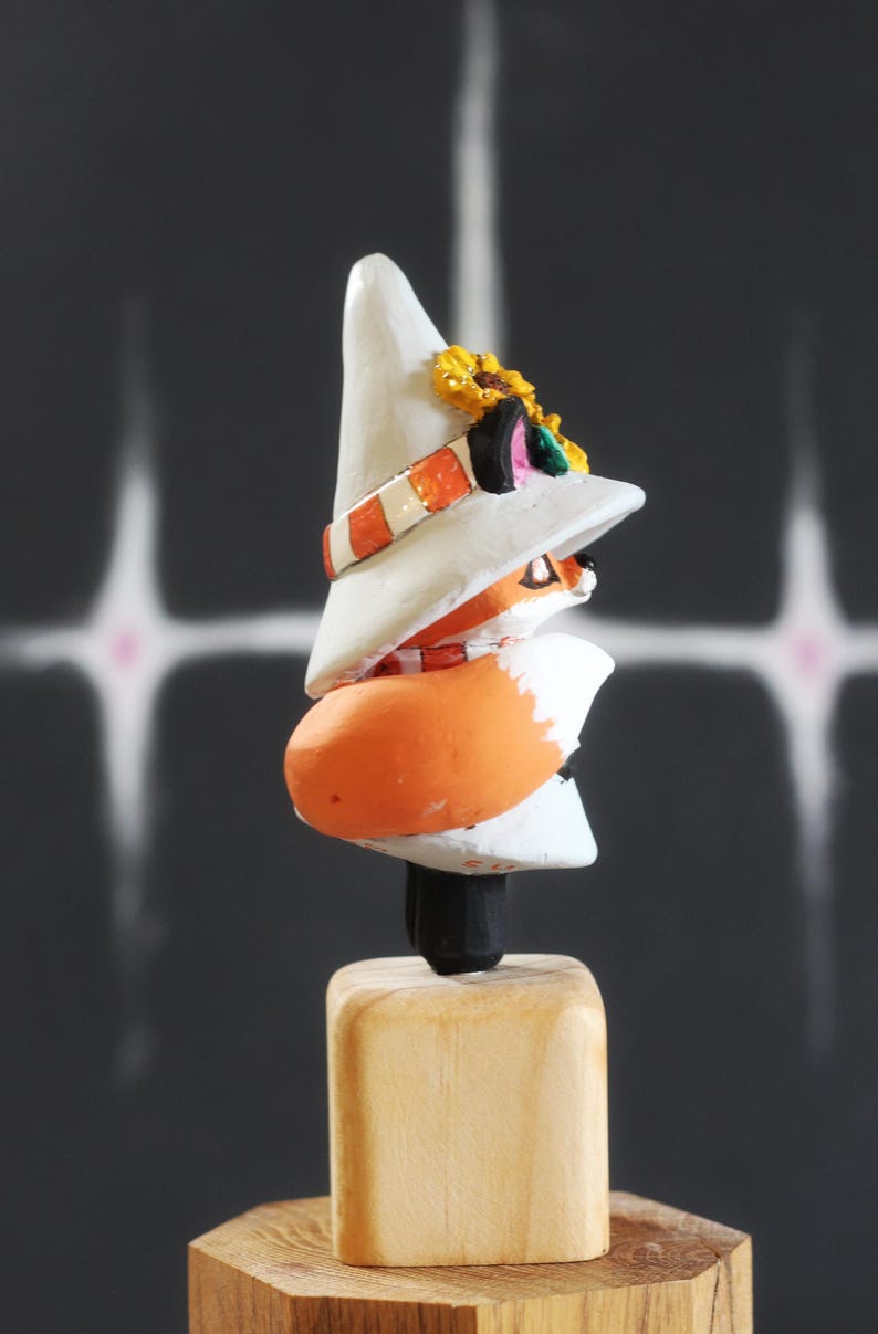 May include: A quirky wooden fox ornament. The fox is depicted wearing a white hat adorned with a sunflower and an orange and white striped band. The fox has an orange body and is mounted on a wooden base. The background is dark with white streaks.
