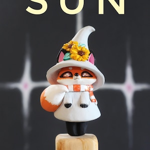 May include: A charming fox figurine, dressed in a white witch hat with yellow sunflowers, a white dress, and an orange and white scarf. The word "SUN" is displayed above the figurine against a dark background.