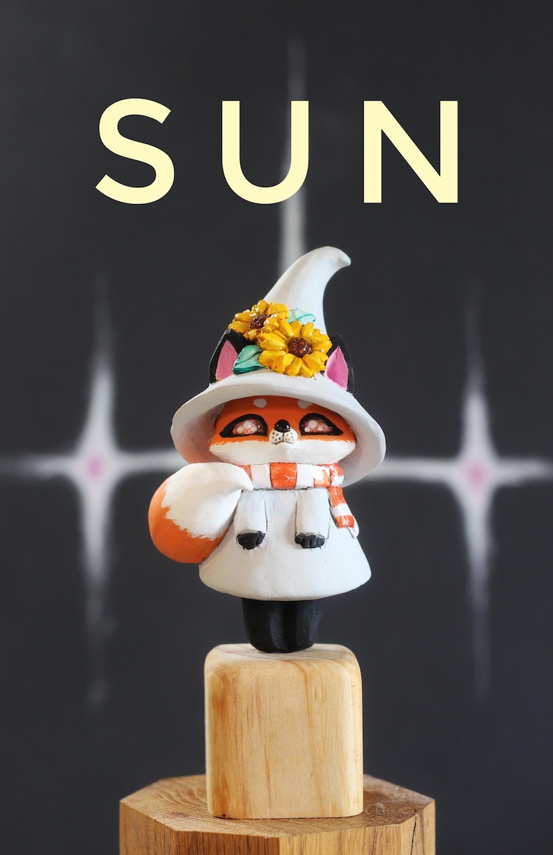 May include: A charming fox figurine, dressed in a white witch hat with yellow sunflowers, a white dress, and an orange and white scarf. The word "SUN" is displayed above the figurine against a dark background.