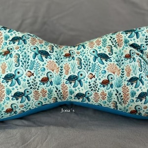 Könnte beinhalten: Ein knochenförmiges Kissen mit hellblauem Hintergrund und einem sich wiederholenden Muster aus Meeresschildkröten, Seepferdchen, kleinen Fischen und Korallen in Blau-, Orange- und Grüntönen. Das Wort Jona's ist in kleiner weißer Schrift vorne mittig aufgedruckt.