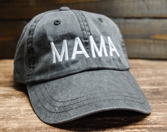 May include: A gray baseball cap with the word "MAMA" embroidered in white. The cap has a curved brim and is made of a soft, worn fabric. The background is a wooden surface.