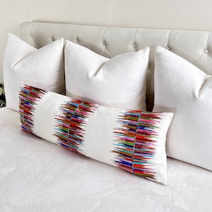 May include: A white bed with a white headboard and three white pillows. The center pillow has a colorful geometric pattern with red, blue, green, and yellow accents.