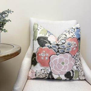 May include: A decorative throw pillow with a floral pattern in shades of pink, red, blue, green, and black. The pillow is placed on a white upholstered chair, creating a cozy and inviting aesthetic. The pillow's design features large, stylized flowers.