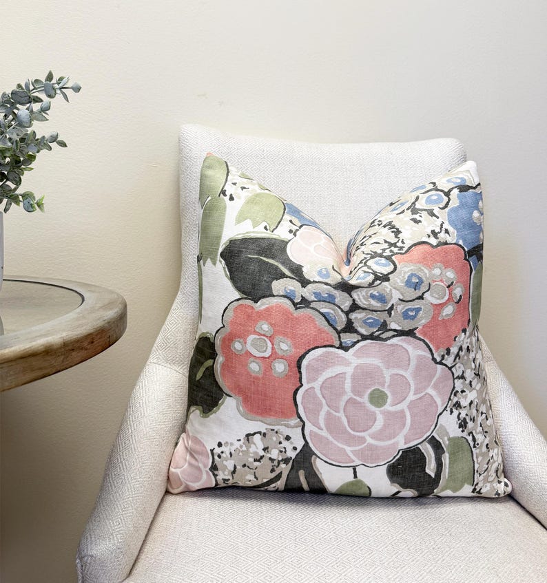May include: A decorative throw pillow with a floral pattern in shades of pink, red, blue, green, and black. The pillow is placed on a white upholstered chair, creating a cozy and inviting aesthetic. The pillow's design features large, stylized flowers.