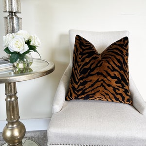 May include: A decorative throw pillow with a tiger stripe pattern in shades of brown and black. The pillow is placed on a cream-colored chair. The room also features a silver side table with a glass top and a bouquet of white roses.