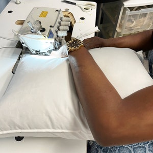 May include: A white pillow being sewn on an industrial sewing machine. The machine is silver and white, with a bright light illuminating the needle and fabric. The person's arm and hand are visible, wearing a gold watch and a white glove.