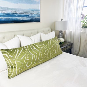 May include: A green and white patterned lumbar pillow on a white bed with white pillows. The bed is in a bedroom with a painting on the wall and a lamp on a side table.