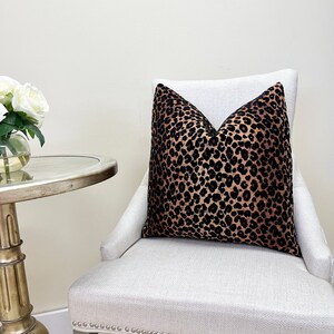 Puede incluir: Un coj&iacute;n de tiro de estampado de leopardo negro y marr&oacute;n sobre una silla blanca con un fondo blanco.