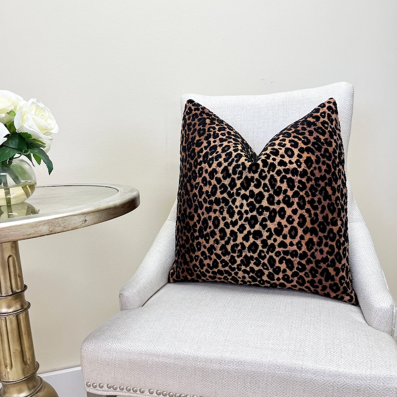 Puede incluir: Un coj&iacute;n de tiro de estampado de leopardo negro y marr&oacute;n sobre una silla blanca con un fondo blanco.