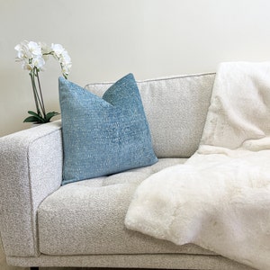 May include: A light blue textured cushion sits on a light grey sofa, beside a white faux fur throw. A white orchid plant in a white pot is on the left. The sofa has a tufted design.