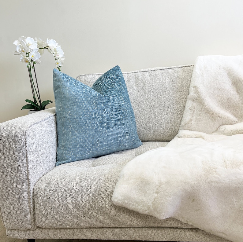 May include: A light blue textured cushion sits on a light grey sofa, beside a white faux fur throw. A white orchid plant in a white pot is on the left. The sofa has a tufted design.