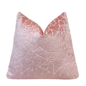 May include: A blush pink decorative pillow with a geometric pattern. The pillow has a soft, velvety texture and a triangular design. The color gradient transitions from a lighter pink to a darker shade.