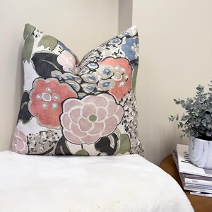 May include: Decorative throw pillow with a floral pattern in shades of pink, coral, blue, and gray on a white background. The pillow features large, stylized flower designs and is placed on a white surface.