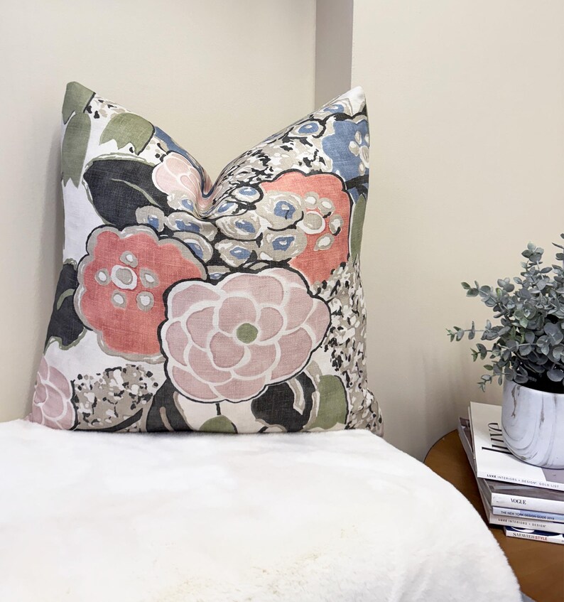 May include: Decorative throw pillow with a floral pattern in shades of pink, coral, blue, and gray on a white background. The pillow features large, stylized flower designs and is placed on a white surface.