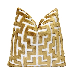 May include: A decorative pillow with a gold and white geometric pattern. The pillow has a soft, textured appearance and a square shape. The design features a repeating maze-like motif in gold against a white background, creating a striking contrast.