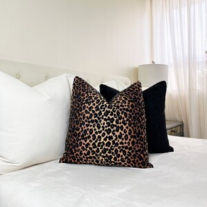 Puede incluir: Una cama blanca con una cabecera blanca y una almohada de estampado de leopardo negro y dorado. La cama est&aacute; hecha con s&aacute;banas blancas y una manta blanca. Tambi&eacute;n hay una almohada negra en la cama.