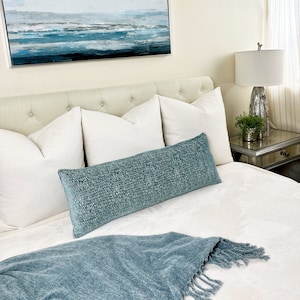 May include: A bedroom setting with a buttoned headboard, white cushions, and a long, textured teal bolster cushion. A matching teal throw with a fringe is at the foot of the bed. An abstract seascape painting hangs above the bed, and a lamp sits on a mirrored bedside table.