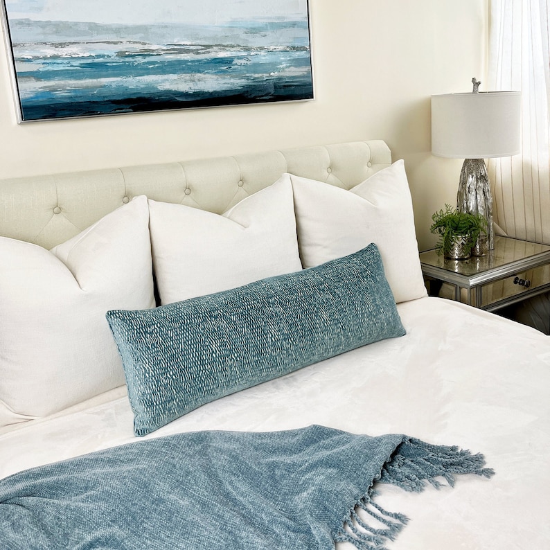 May include: A bedroom setting with a buttoned headboard, white cushions, and a long, textured teal bolster cushion. A matching teal throw with a fringe is at the foot of the bed. An abstract seascape painting hangs above the bed, and a lamp sits on a mirrored bedside table.