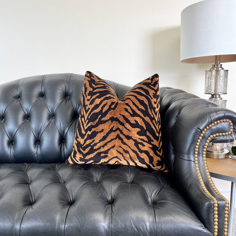 May include: A decorative throw pillow with a tiger stripe pattern in shades of brown and black. The pillow is placed on a black leather sofa with button tufting and gold-colored nailhead trim. The pillow adds a touch of animal print to the room's decor.