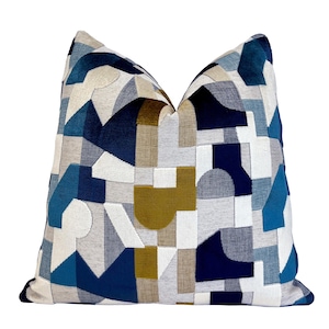May include: Decorative throw pillow with a geometric pattern in shades of blue, gray, white, and olive green. The pillow features a patchwork design with various shapes and textures, creating a modern aesthetic.
