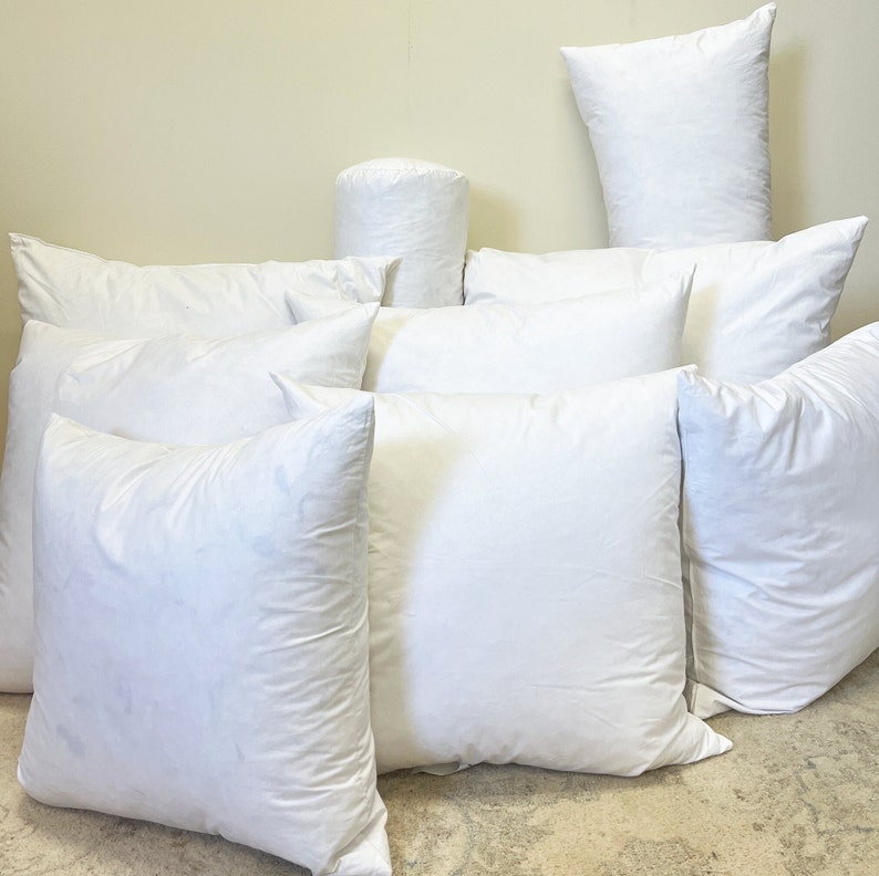 May include: A collection of white pillows in various shapes and sizes. The assortment includes square, rectangular, and cylindrical pillows, all with a soft, puffy appearance. The pillows are arranged in a casual, layered composition, suggesting a focus on comfort and bedding.