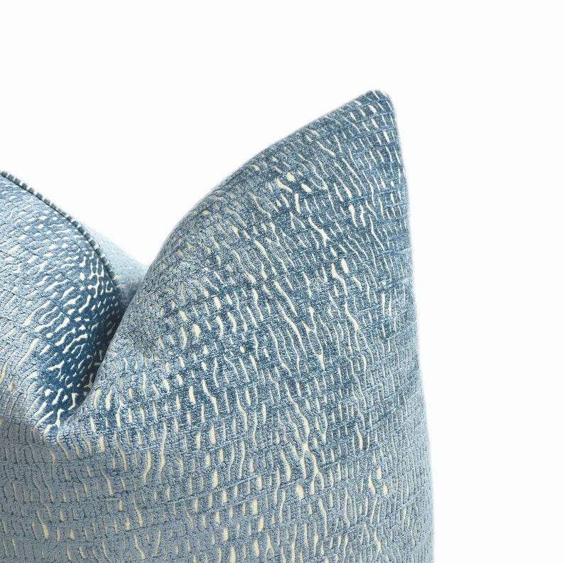 May include: Close-up of a blue decorative cushion with a textured pattern. The cushion has a light blue background with a cream-coloured, vertical, wavy pattern. The cushion is made of a soft fabric and is a decorative home accent.