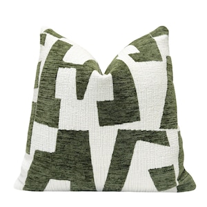 May include: A decorative square pillow with a geometric pattern. The pillow features a textured, olive green fabric with large white shapes. The design creates a bold, modern look, suitable for home decor.