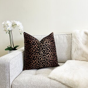 Puede incluir: Un coj&iacute;n de sof&aacute; de color marr&oacute;n y negro con estampado de leopardo con una manta de piel sint&eacute;tica blanca sobre un sof&aacute; gris claro.