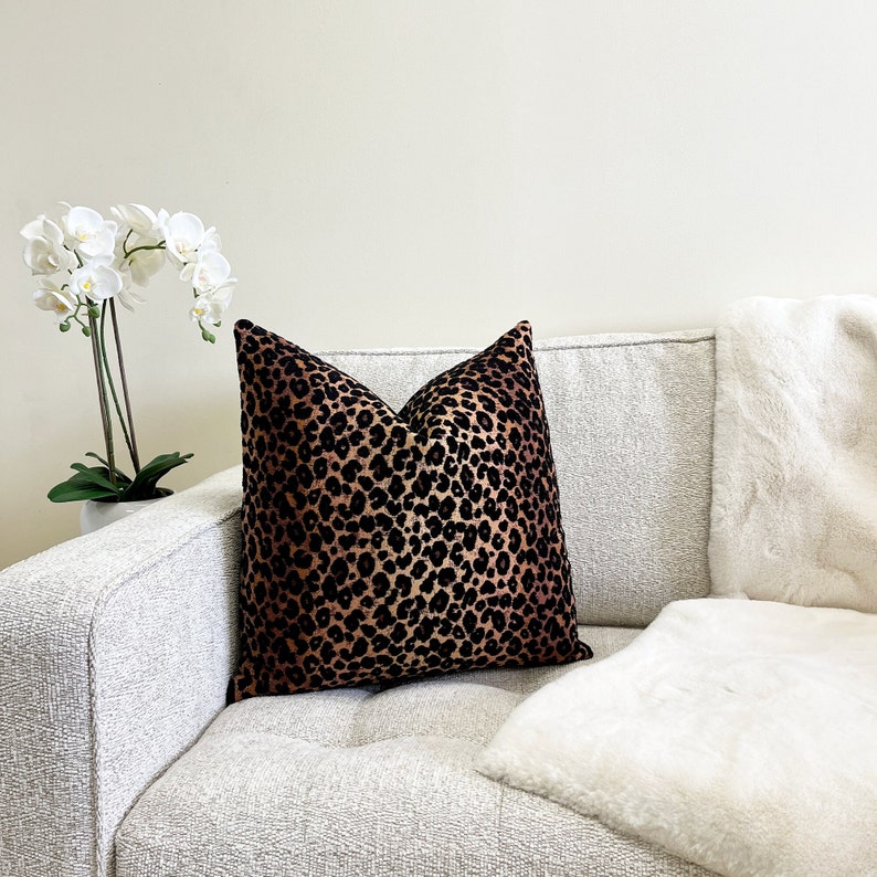 Puede incluir: Un coj&iacute;n de sof&aacute; de color marr&oacute;n y negro con estampado de leopardo con una manta de piel sint&eacute;tica blanca sobre un sof&aacute; gris claro.