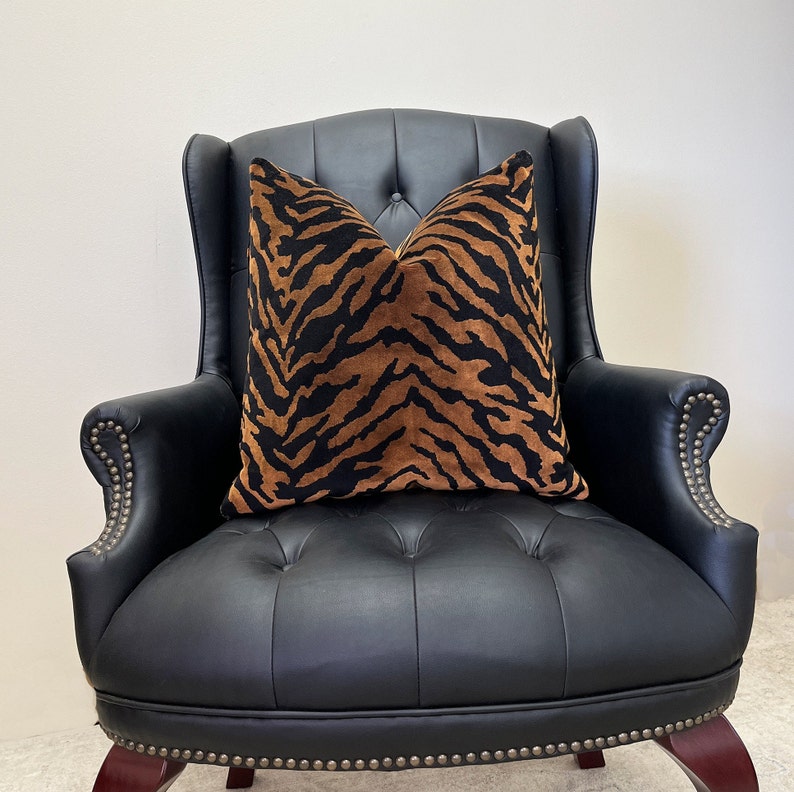 May include: A decorative throw pillow with a tiger stripe pattern in black and brown sits on a black leather armchair. The pillow has a soft, textured appearance, and the armchair features a button-tufted design.