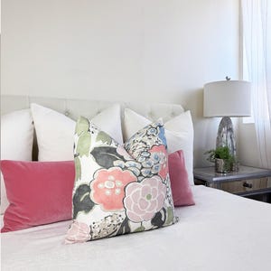 May include: A bed with several pillows. The pillows include white, pink, and floral patterned designs. The floral pillow has pink, blue, and green flowers. A white lamp with a gray base sits on a mirrored nightstand.