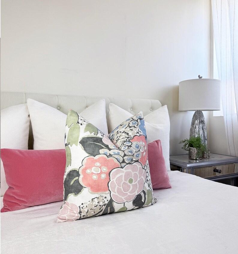 May include: A bed with several pillows. The pillows include white, pink, and floral patterned designs. The floral pillow has pink, blue, and green flowers. A white lamp with a gray base sits on a mirrored nightstand.