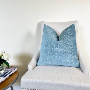 May include: A large, square, blue cushion with a textured pattern sits on a light-coloured upholstered chair. The cushion's colour gradient shifts from a darker blue at the top to a lighter shade at the bottom. The chair has a high back and a patterned seat.