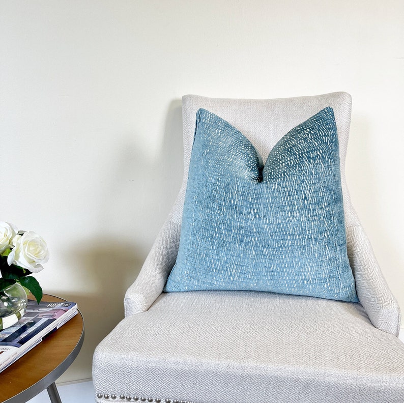 May include: A large, square, blue cushion with a textured pattern sits on a light-coloured upholstered chair. The cushion's colour gradient shifts from a darker blue at the top to a lighter shade at the bottom. The chair has a high back and a patterned seat.