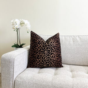Puede incluir: Un sof&aacute; blanco con un coj&iacute;n de estampado de leopardo negro y marr&oacute;n. El sof&aacute; est&aacute; en una habitaci&oacute;n con paredes blancas y una planta de orqu&iacute;dea blanca en una maceta.