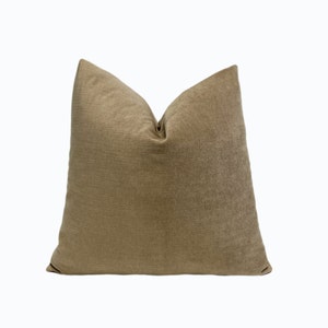 May include: A square decorative pillow in a neutral tan color. The pillow has a textured surface and a soft, inviting appearance. The pillow is designed for home decor and adds a touch of elegance to any living space.