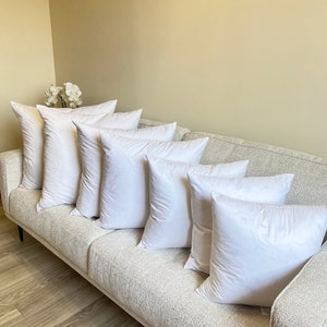 May include: Seven white square pillows arranged on a light-colored textured sofa. The pillows are plump and appear to be filled with a soft material. The sofa is positioned against a light beige wall, creating a neutral and inviting setting.