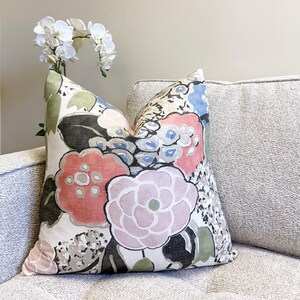 May include: Square decorative pillow with a large floral pattern in shades of pink, red, blue, green, and black on a cream background. The pillow is placed on a light gray sofa. A white orchid is in the background.