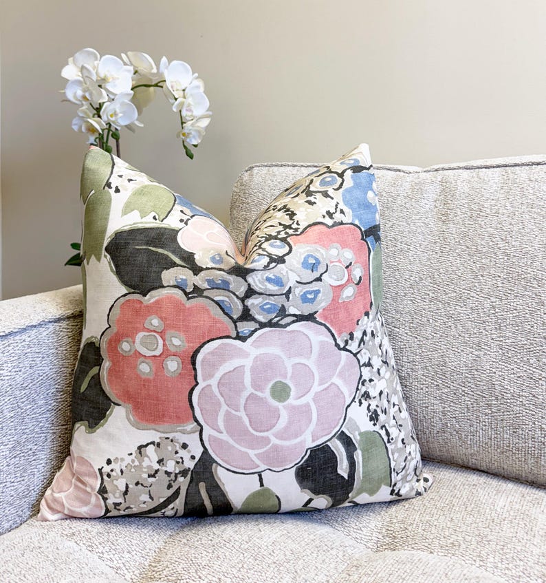 May include: Square decorative pillow with a large floral pattern in shades of pink, red, blue, green, and black on a cream background. The pillow is placed on a light gray sofa. A white orchid is in the background.