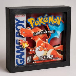 May include: A framed artwork featuring the Pokémon Red Version Game Boy game box art. The artwork showcases a vibrant orange Charizard with blue wings against a red background. The black frame surrounds the game's logo and text, including "Gotta catch 'em all!"